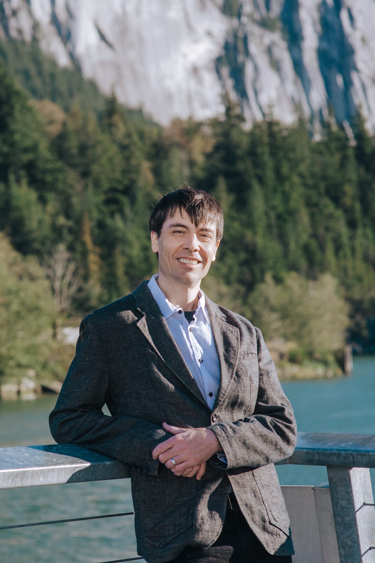 Chris Pettingill in a grey linen blazer, leaning on a railing at the Squamish waterfront with the mountainside behind him