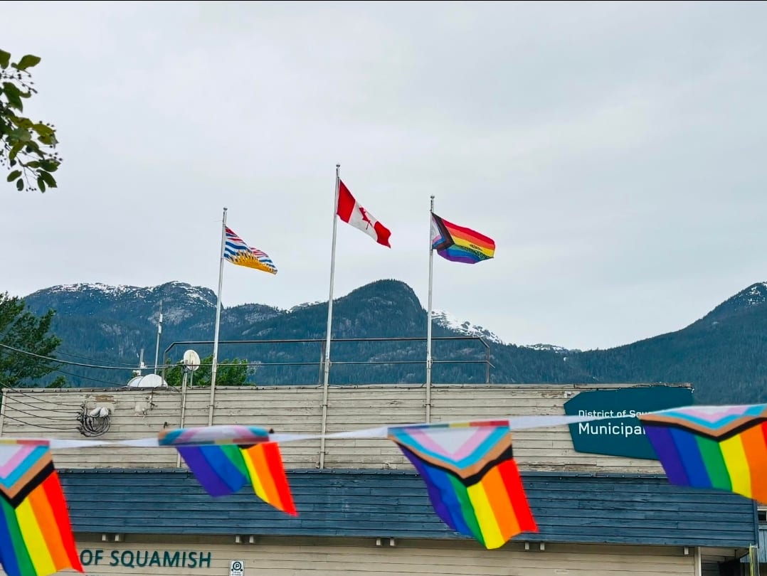 The Canadian, British Columbia and Progress Pride flags flying over the District of Squamish municipal hall, Progress Pride bunting strung in front and the mountains behind