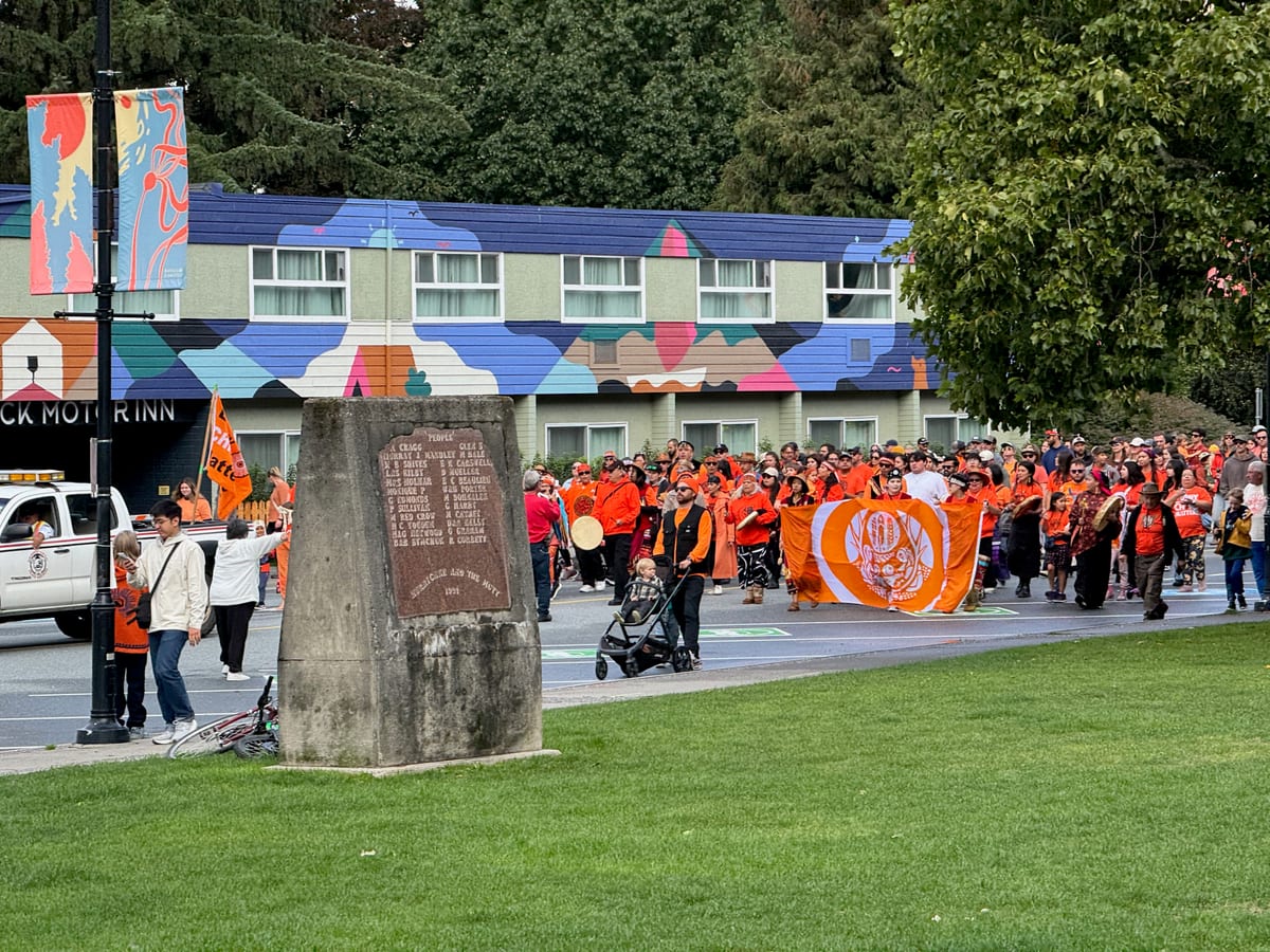 A large group in orange shirts walking through downtown Squamish on Orange Shirt Day, carrying an orange banner and a hand drum