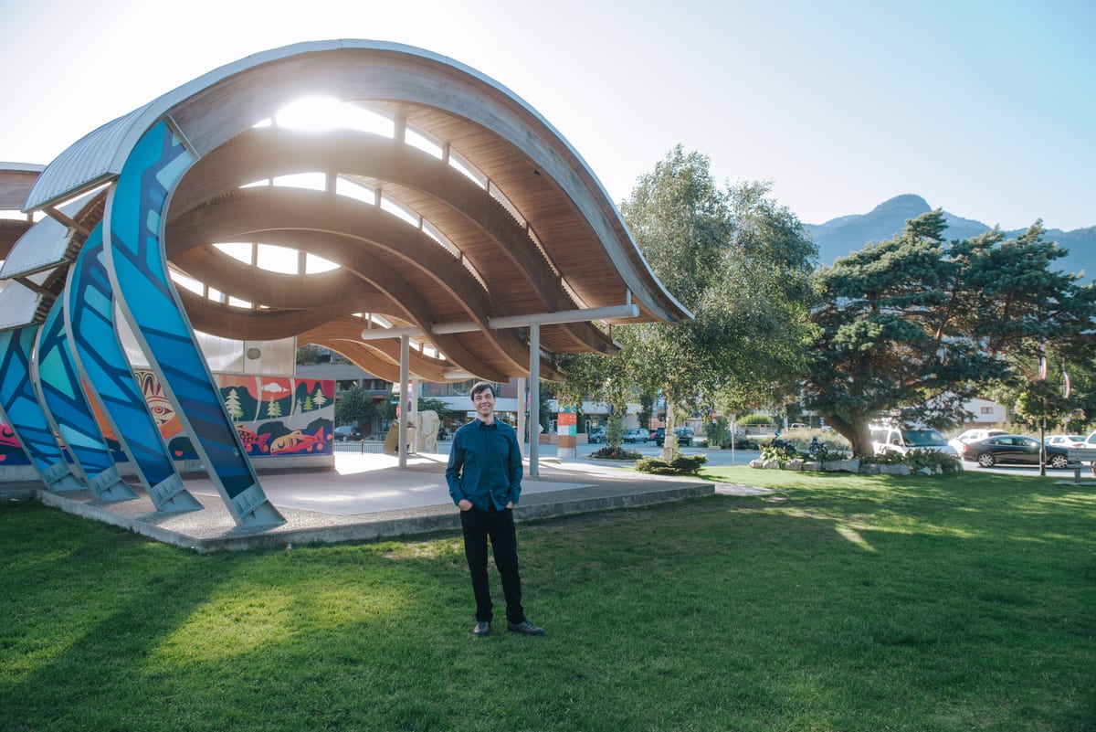Chris Pettingill standing on the lawn in front of the O'Siyam Pavilion in downtown Squamish, the Squamish Nation salmon mural and the mountains behind him
