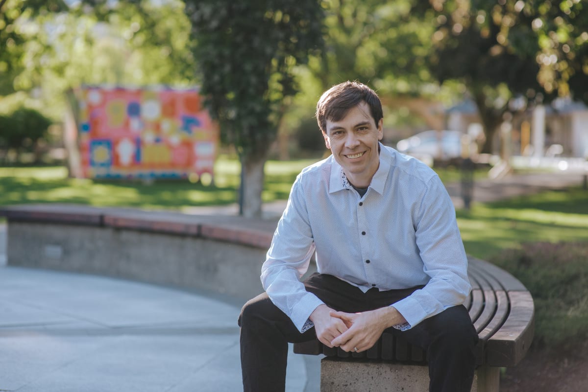Chris Pettingill sitting on a bench in a downtown Squamish park, a bright mural on the grass behind him
